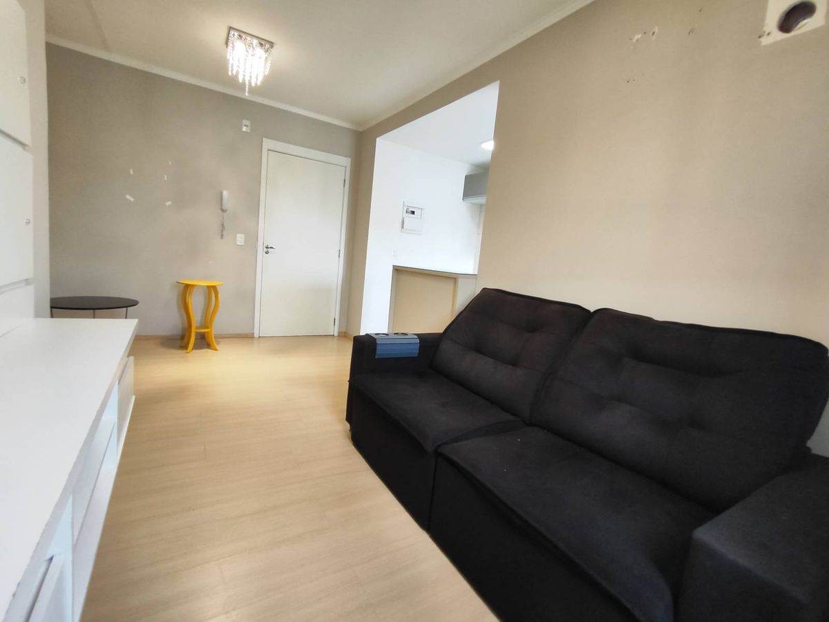 Sala com sofá preto, paredes claras, piso de madeira, iluminação artificial