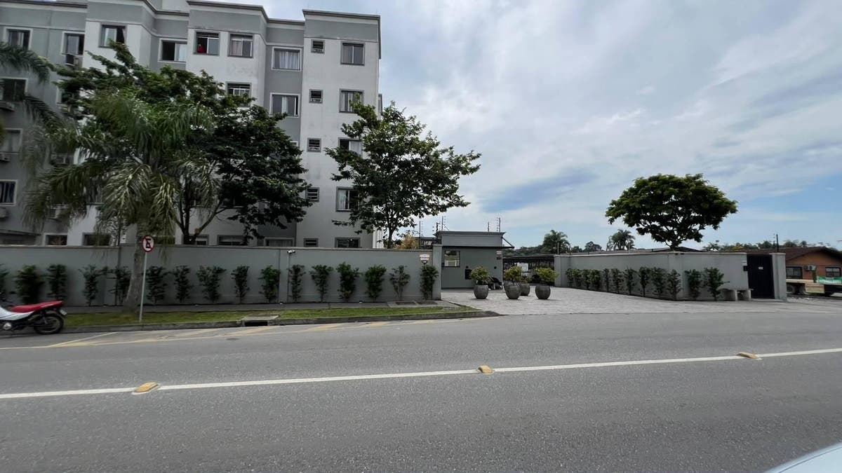 Fachada de edifício com muro e vegetação ao redor, sob céu nublado