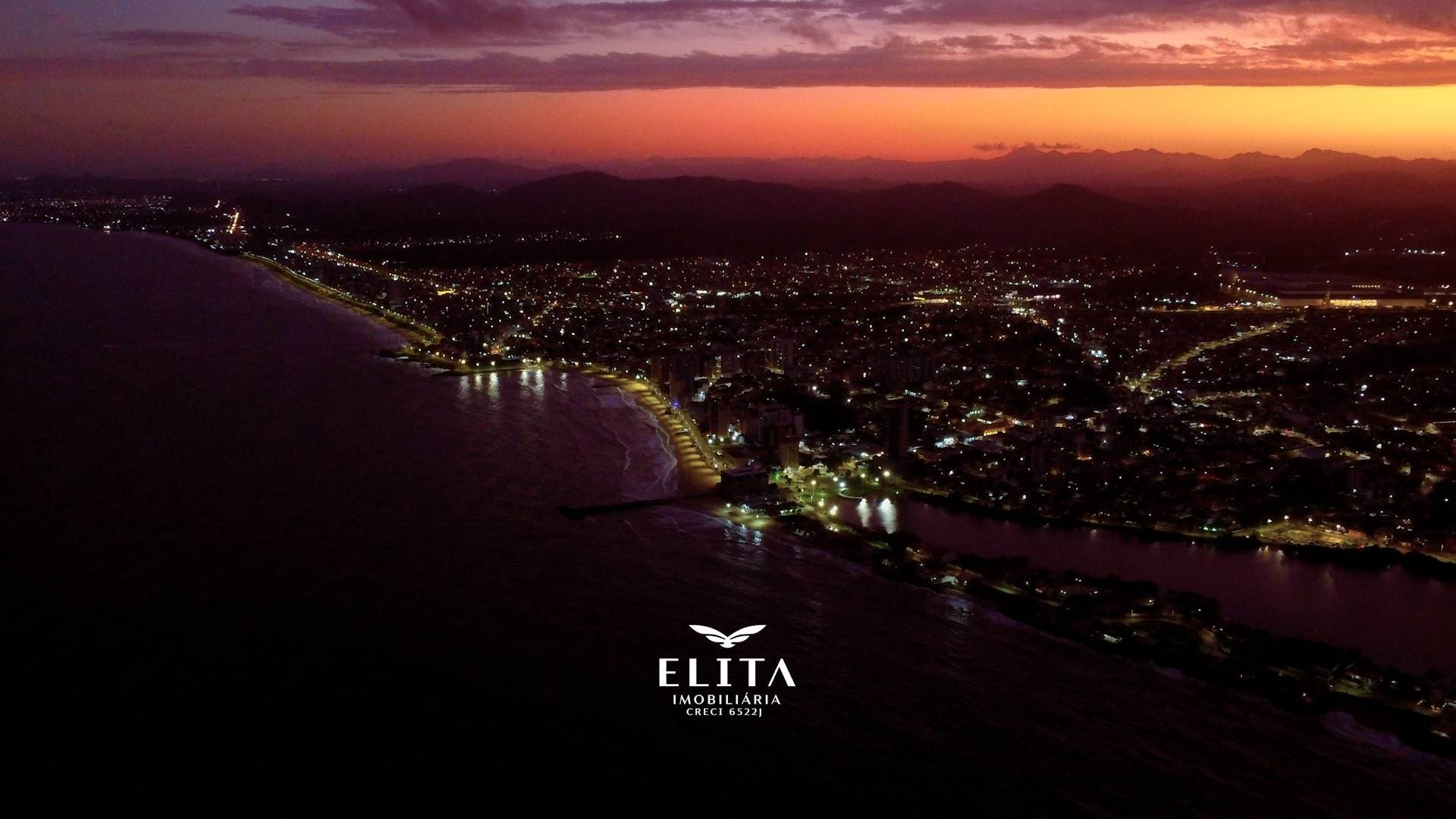 Orla iluminada ao entardecer com reflexos, casas e edifícios de Barra Velha