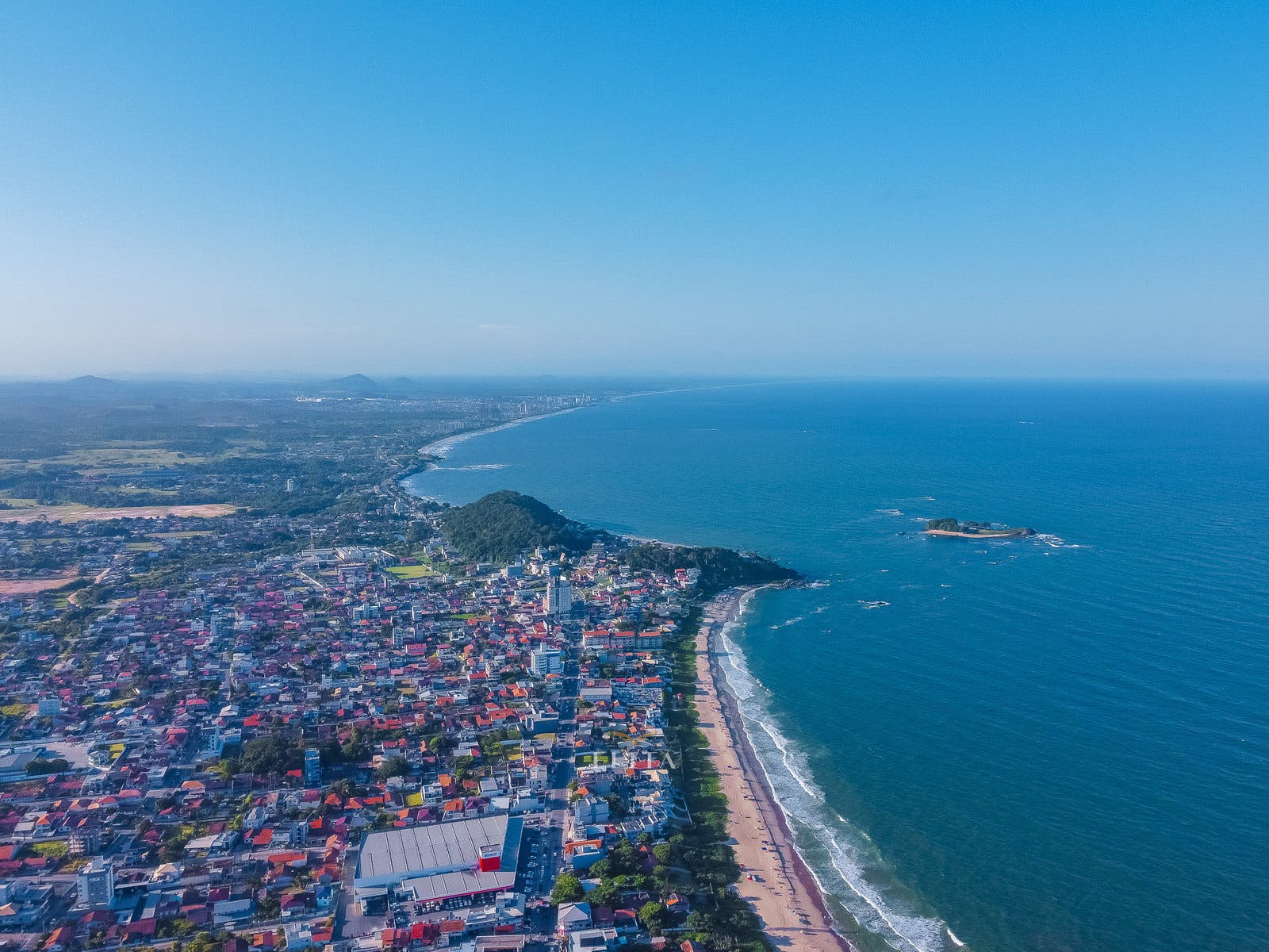 Vista aérea de cidade costeira, praia extensa, morro arborizado e ilha rochosa diante do litoral, sob céu azul.